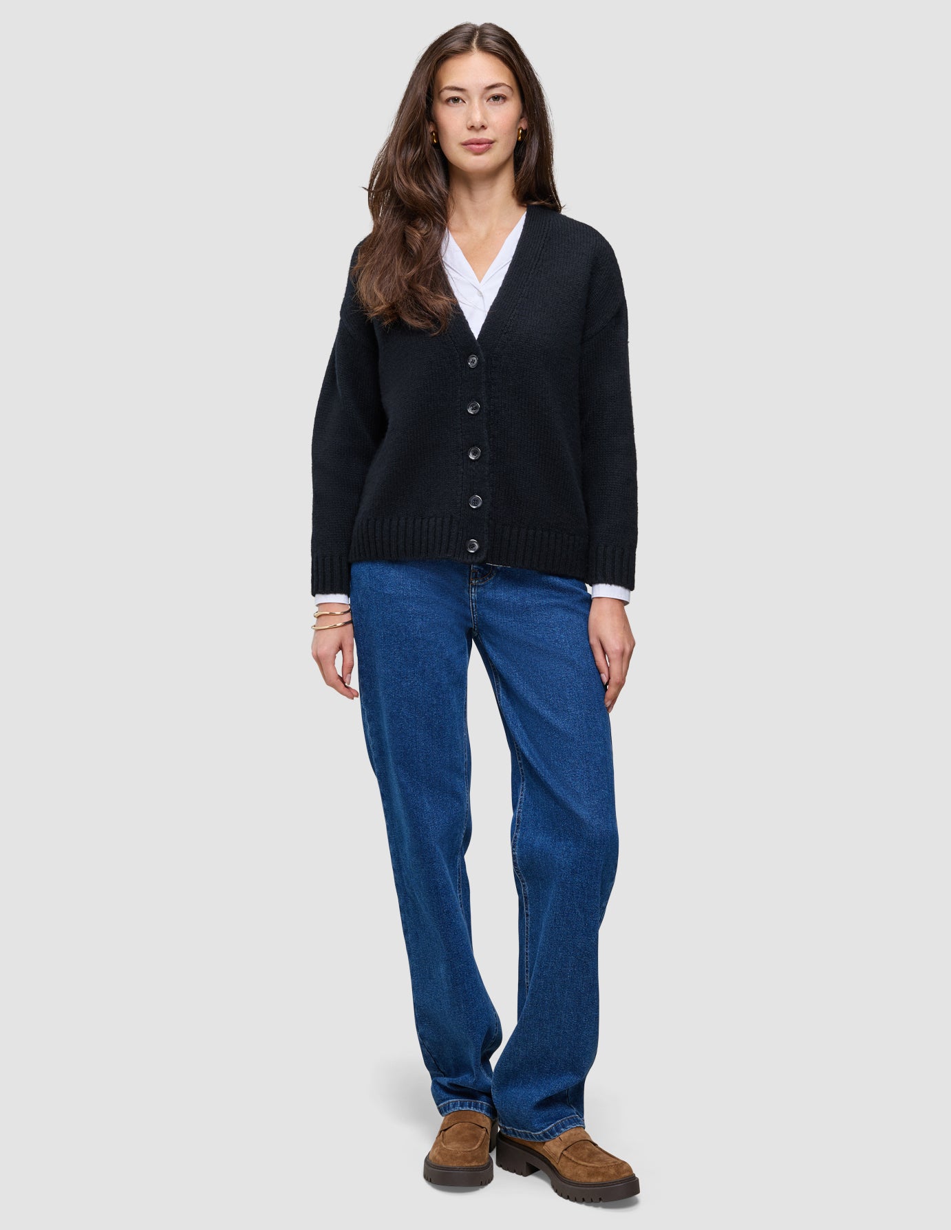 En person bär en svart, tjock och mjuk stickad damkofta i ull över en vit skjorta och blå jeans samt bruna loafers.